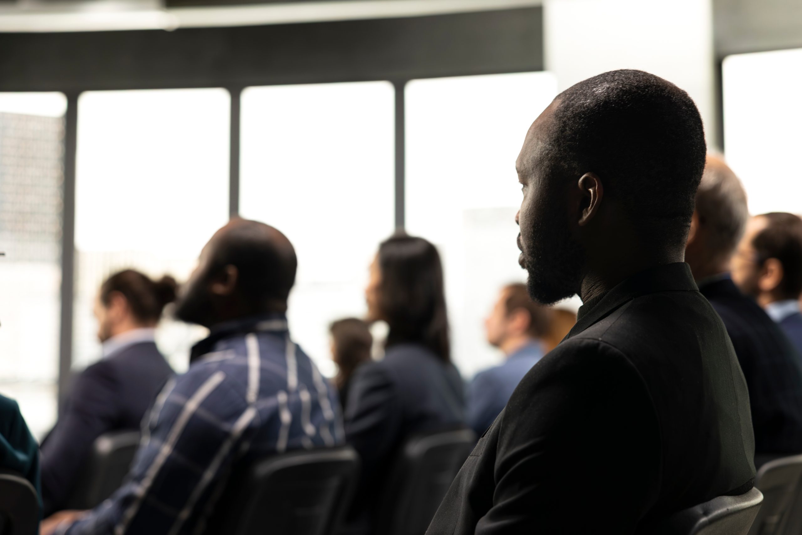 Black male attendee listening to presentation with diverse people in audience, business conference event with presenter. Chairs fill auditorium as participants ... See More