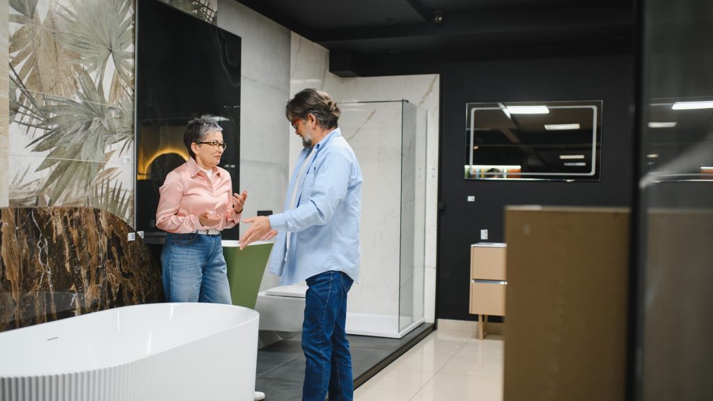 Designer and homeowner reviewing a measured bathroom floor plan beside a tiled vanity display in a kitchen and bath showroom