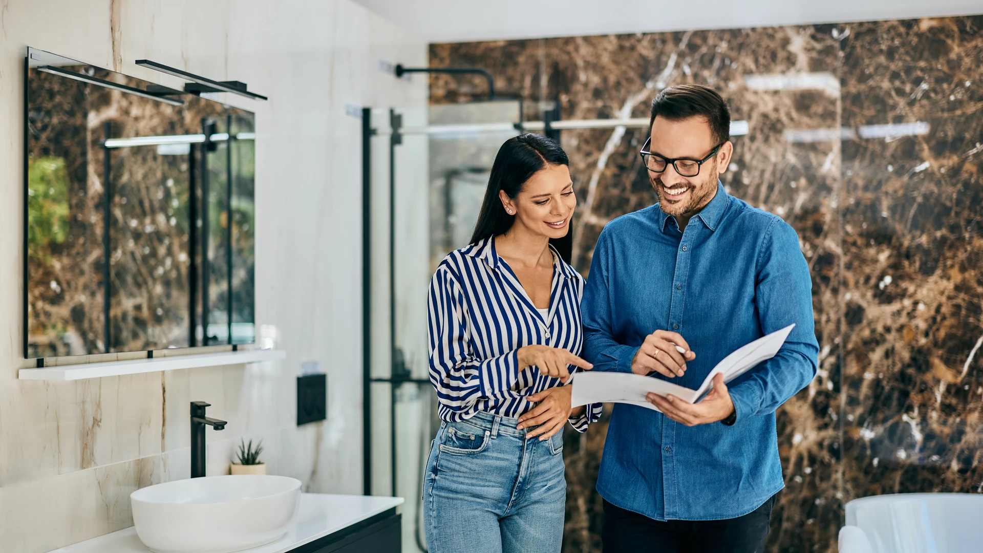 Two people reviewing documents in a bathroom with a vessel sink and dark veined wall