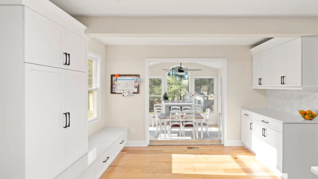Bright white kitchen with a built-in window bench and tall cabinets opening into a sunroom dining area with an arched window and trees outside