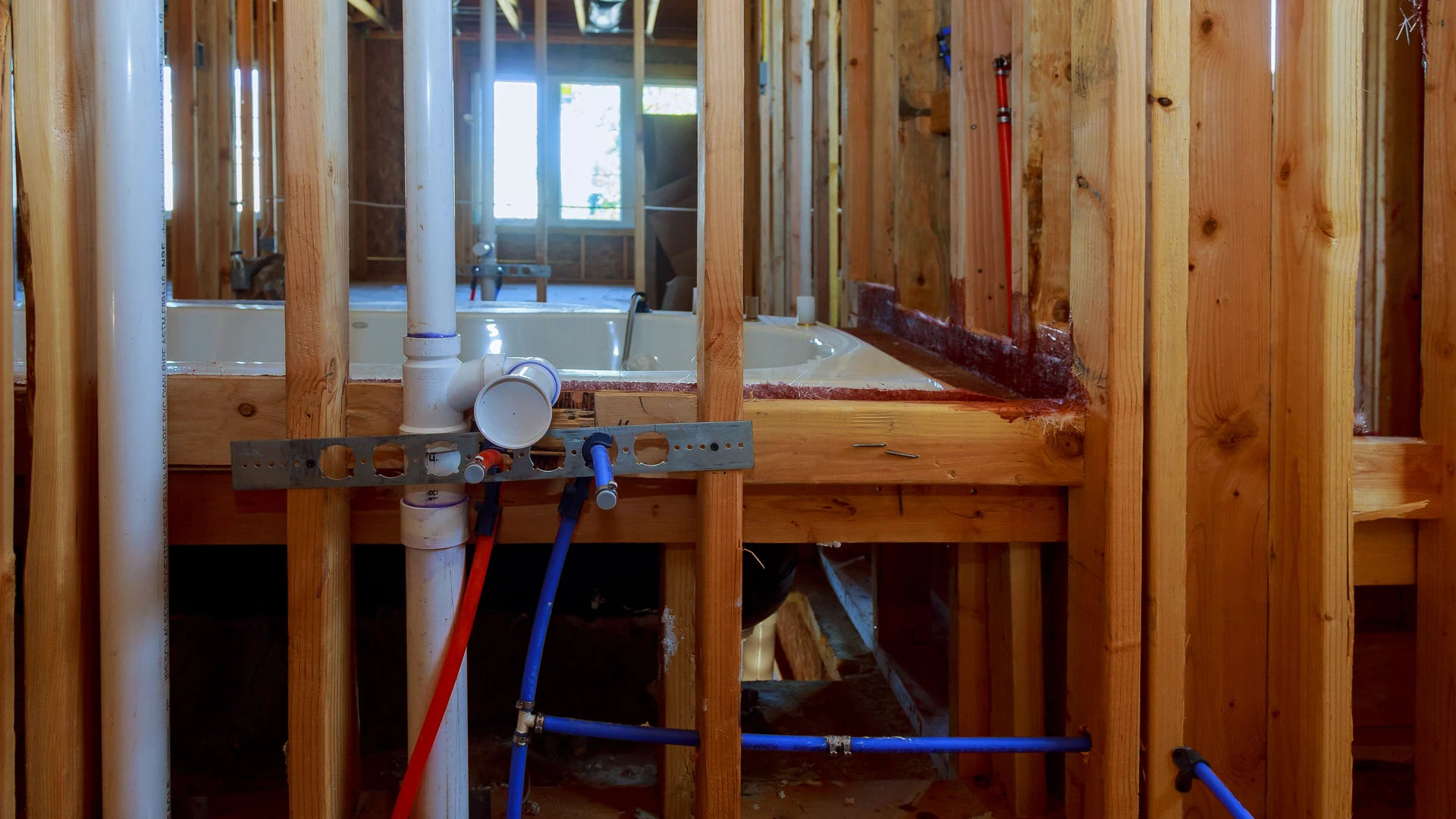Bathroom plumbing rough-in with a white drain pipe and red and blue supply lines in open wall framing