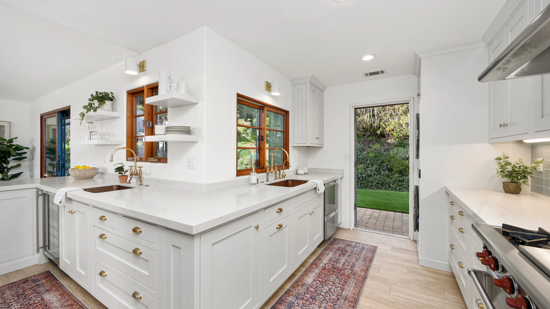 White shaker kitchen with banks of drawers, open shelves, gold-colored fixtures and brown window frames.