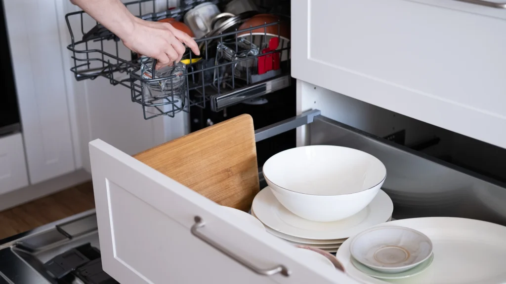 Open dishwasher rack being loaded with dishes and glassware, beside its own closed white panel-front door that matches the surrounding kitchen cabinets