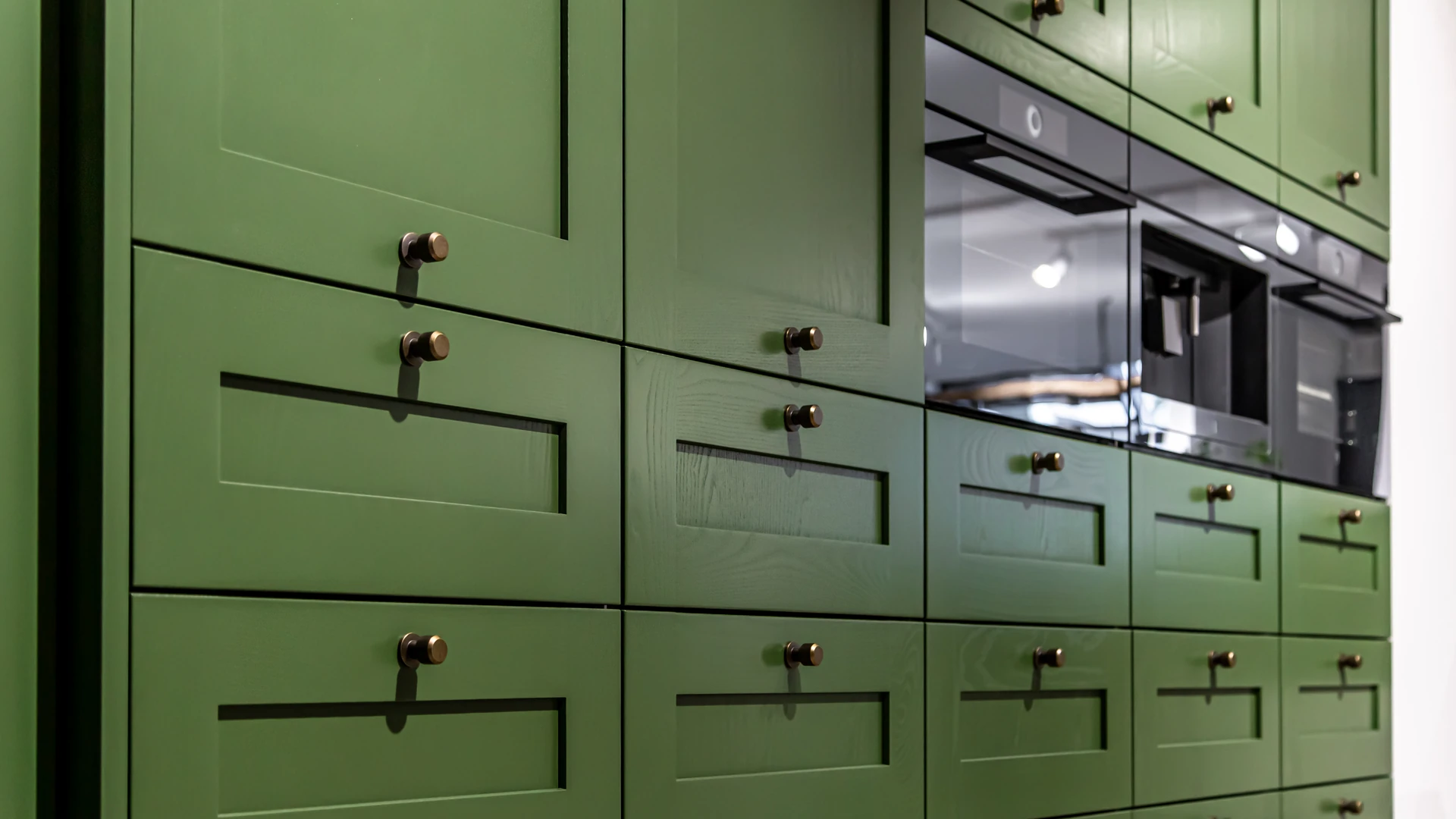Wall of green recessed-panel cabinet doors and drawers with dark knobs around built-in ovens