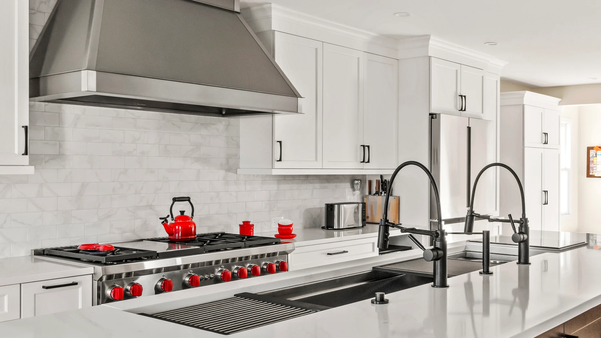 Range with red knobs under a large metallic hood, with two tall island faucets in the foreground.