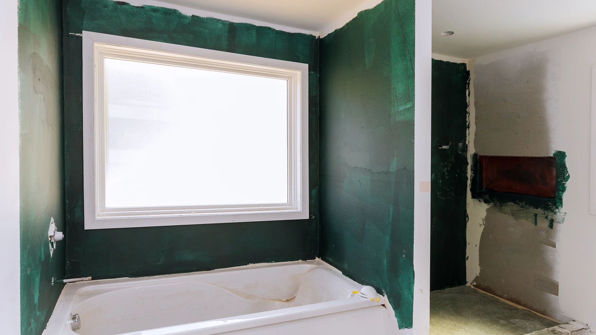 Unfinished bathroom with green-coated walls around a bathtub and a large window