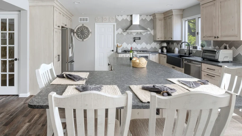 Dark gray speckled countertop on a large island with white slat-back chairs and place mats along its near edge, facing pale cabinets, a dark farmhouse sink and a diamond-pattern tile backsplash
