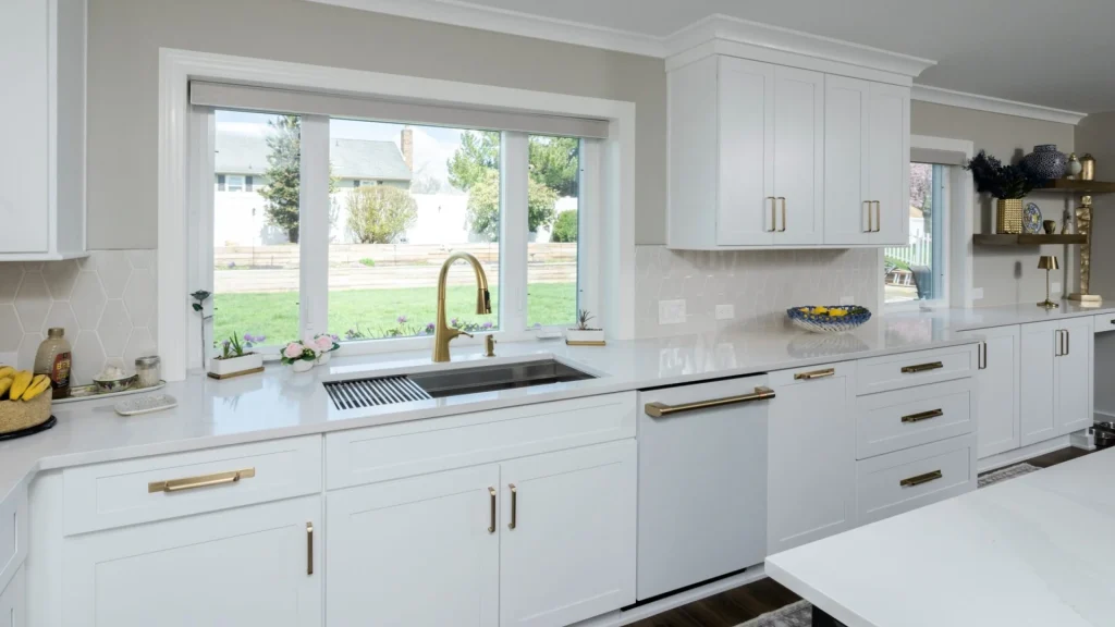 White kitchen with a stainless sink under a wide window, a brass-toned pull-down faucet, a white dishwasher beside the sink, white shaker cabinets with brass-toned pulls, pale hexagon tile and wall outlets above the counter
