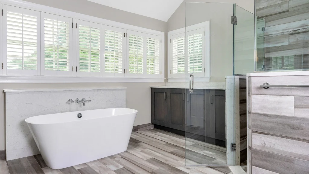 White freestanding tub with a wall-mounted faucet beneath shuttered windows, beside a glass shower and dark vanity cabinets on a wood-look floor