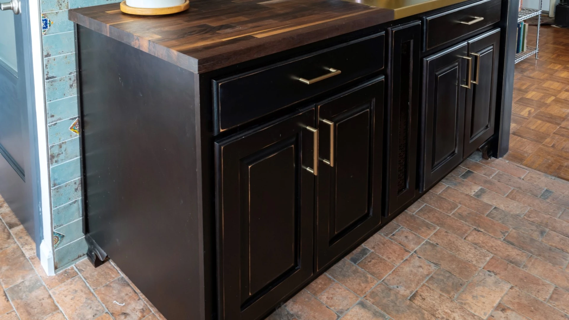 Dark brown base cabinets with raised-panel doors and drawers, each with a gold-tone bar pull, under a dark wood-grain counter on a brick-pattern tile floor