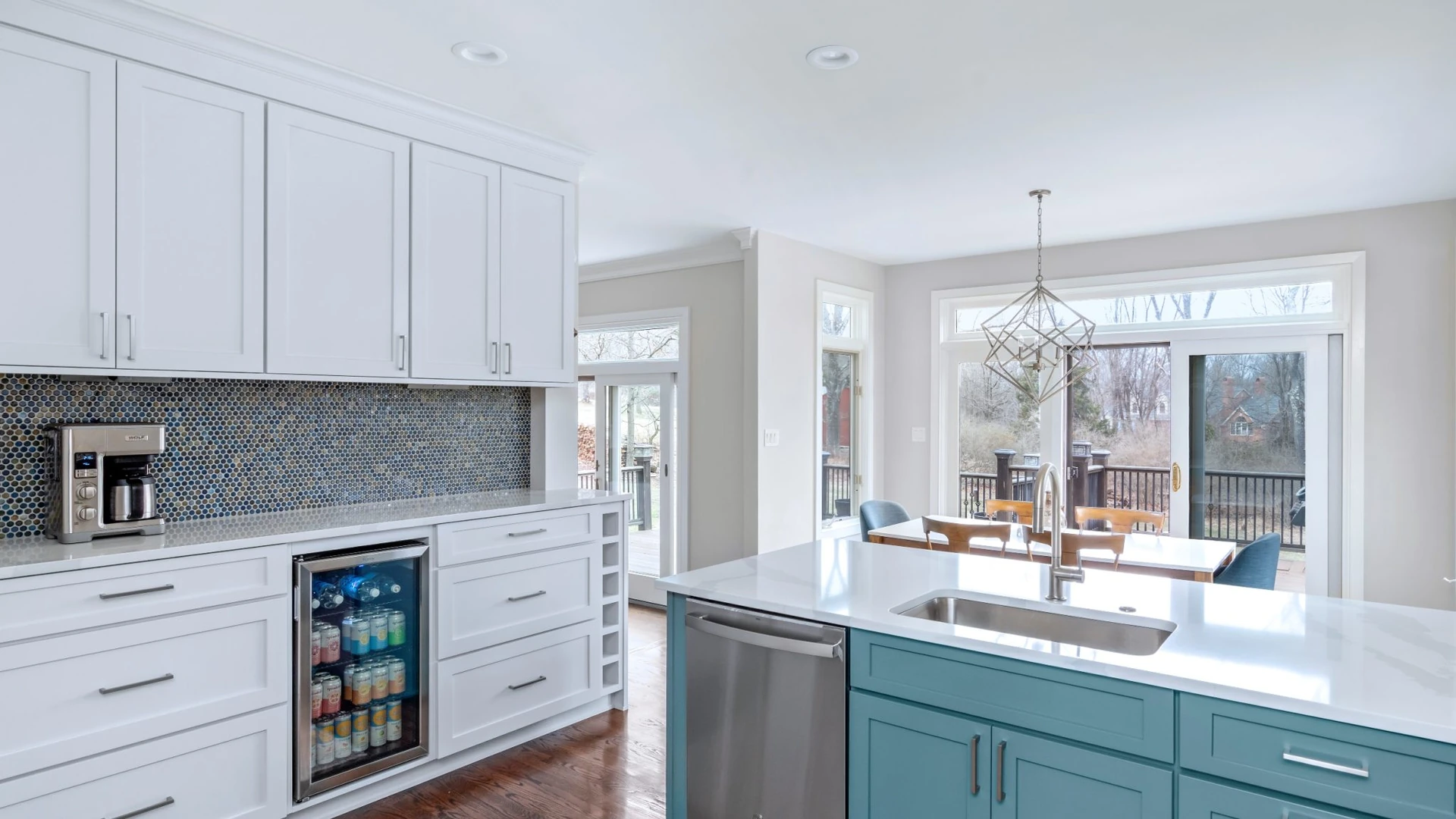 Teal kitchen base cabinets with a white countertop, an undermount stainless sink with a tall faucet and a stainless dishwasher at the sink end, beside white cabinets with a beverage refrigerator and a dining area with sliding glass doors beyond
