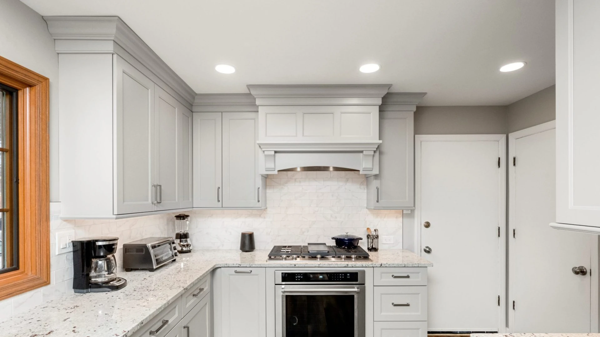 Light gray kitchen with a speckled white countertop that turns an inside corner and continues to a gas cooktop under a paneled hood, with a white veined tile backsplash and a wall oven below