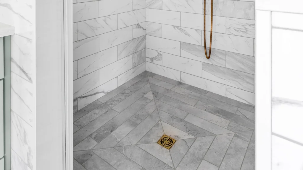 Gray herringbone-tile shower floor with a gold-tone square drain, below white veined wall tile and the end of a hand-held shower hose