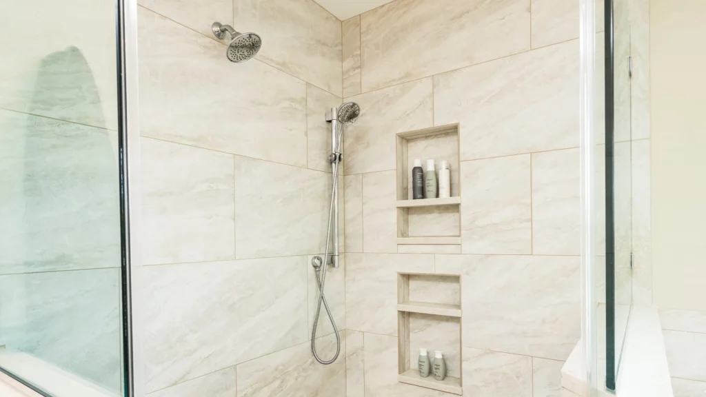 Tiled shower corner with a wall-mounted showerhead, a hand-held shower on a slide bar with its hose, and two tall recessed niches holding shampoo bottles