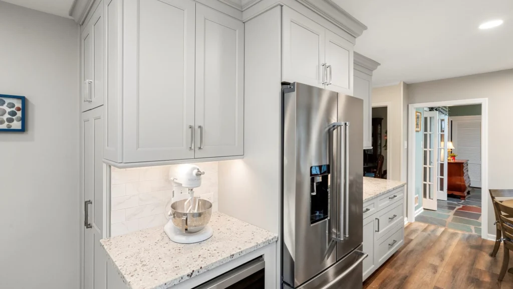 A white stand mixer on a speckled granite counter under lit gray upper cabinets, beside a stainless steel refrigerator, with a hallway and French doors behind