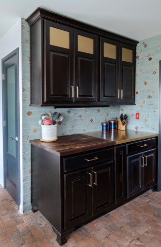 Dark kitchen cabinets with light upper inserts above a counter and blue-green tiled backsplash